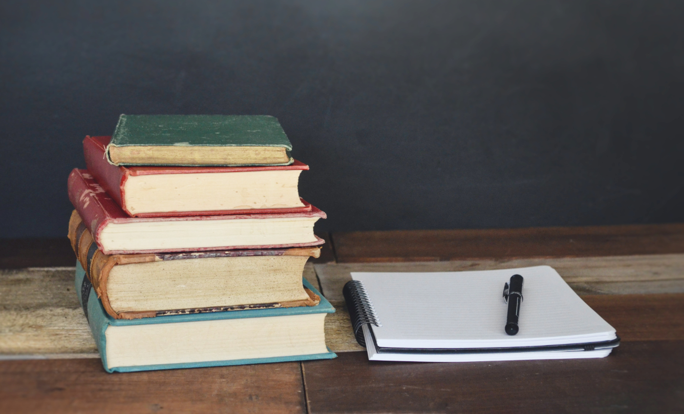 A stack of aged books and a notepad with a pen lying on a table.